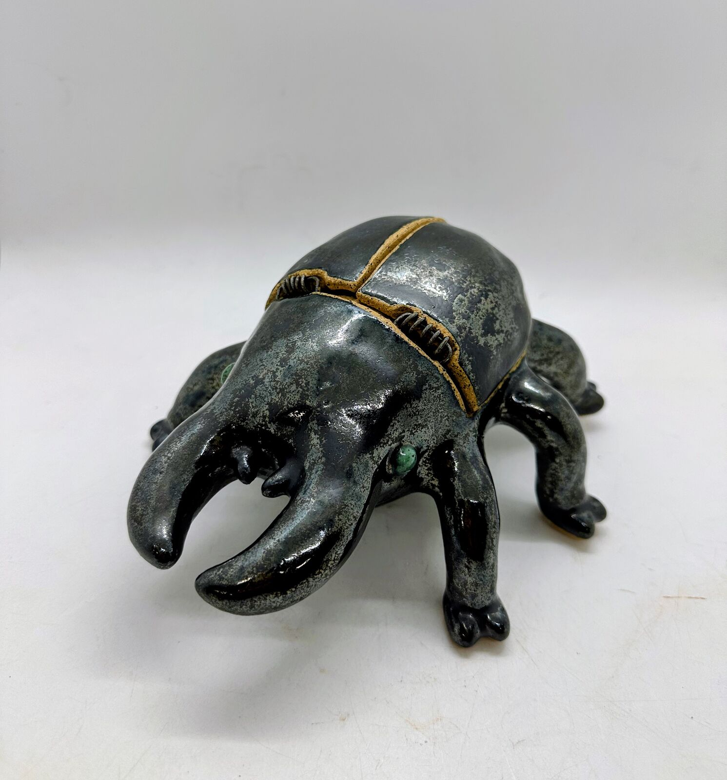 A glazed ceramic stag beetle in speckled gunmetal grey, with curved mandibles, six stubby legs, and green eyes. Its wing-case lid is split down the middle and attached with coiled wire hinges.