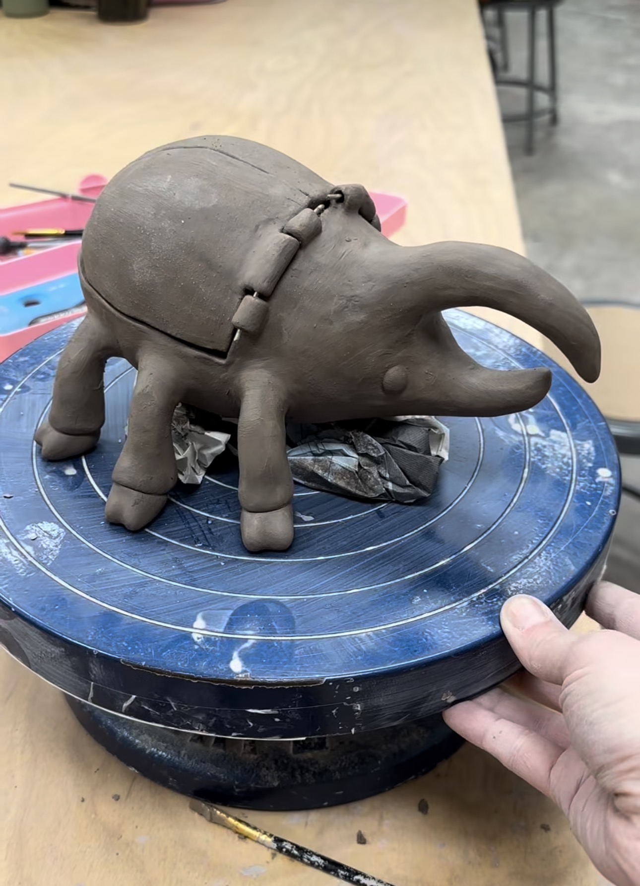 A whimsical horned beetle creature sculpted from grey clay, with a domed shell body, curved horn, and many stubby legs, sitting on a blue banding wheel in the studio.
