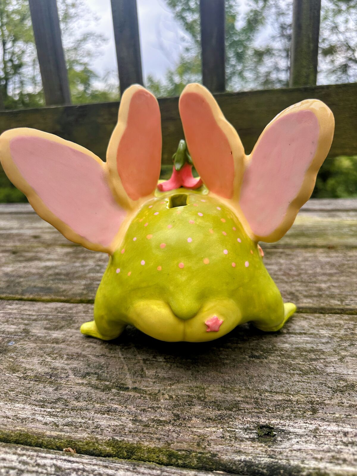 The fairy frog from behind: a round chartreuse body dotted with pink and orange spots, a coin slot on top, a tiny pink star on its bottom, and four butterfly wings standing up like ears.