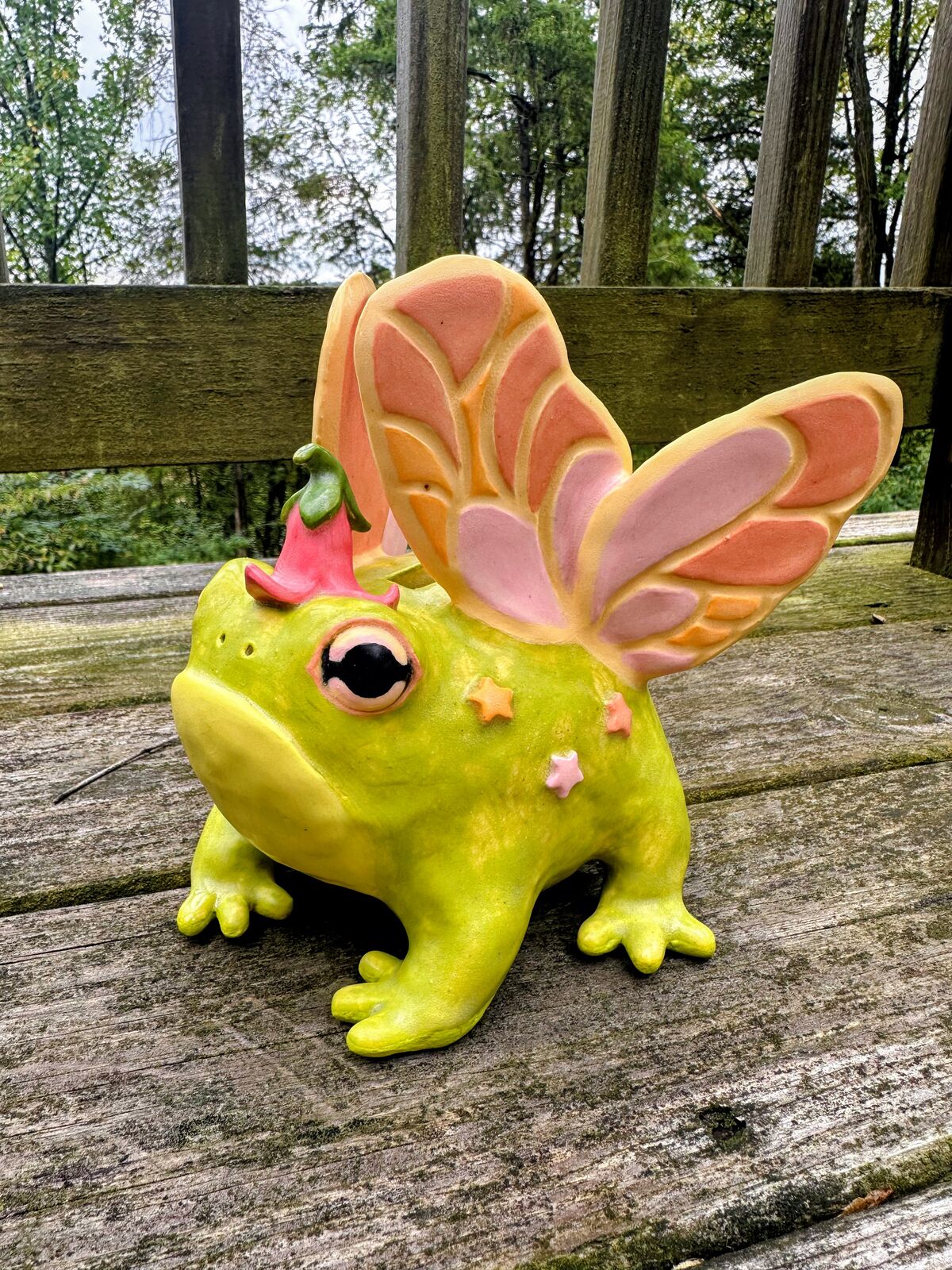 A hand-built ceramic fairy frog in bright chartreuse, with big pink-rimmed eyes, a pink bellflower cap, pastel star spots, and two pairs of pink, peach, and cream butterfly wings, sitting on a weathered wooden deck.