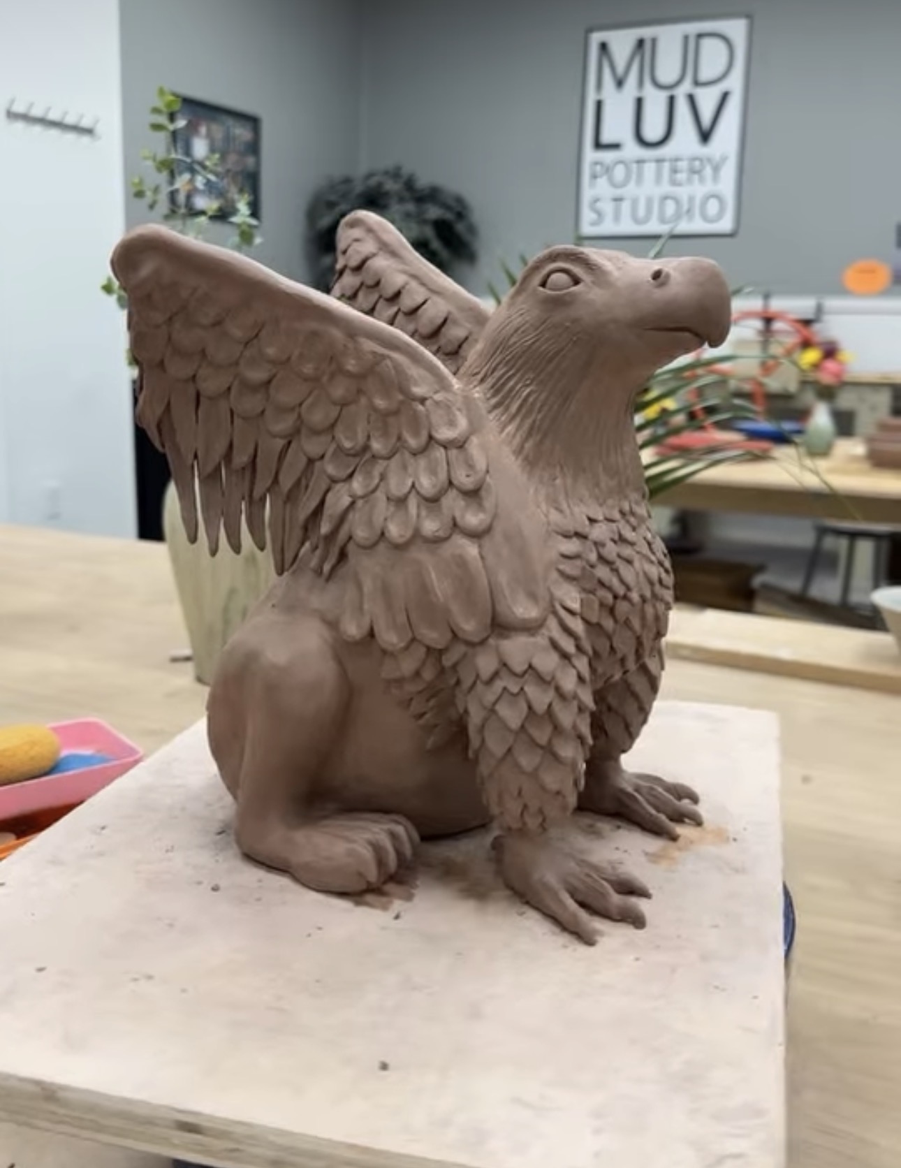 A hand-built raw terracotta clay griffin sculpture with an eagle's head, raised feathered wings, and a lion's body, sitting on a work board in the studio, not yet glazed.